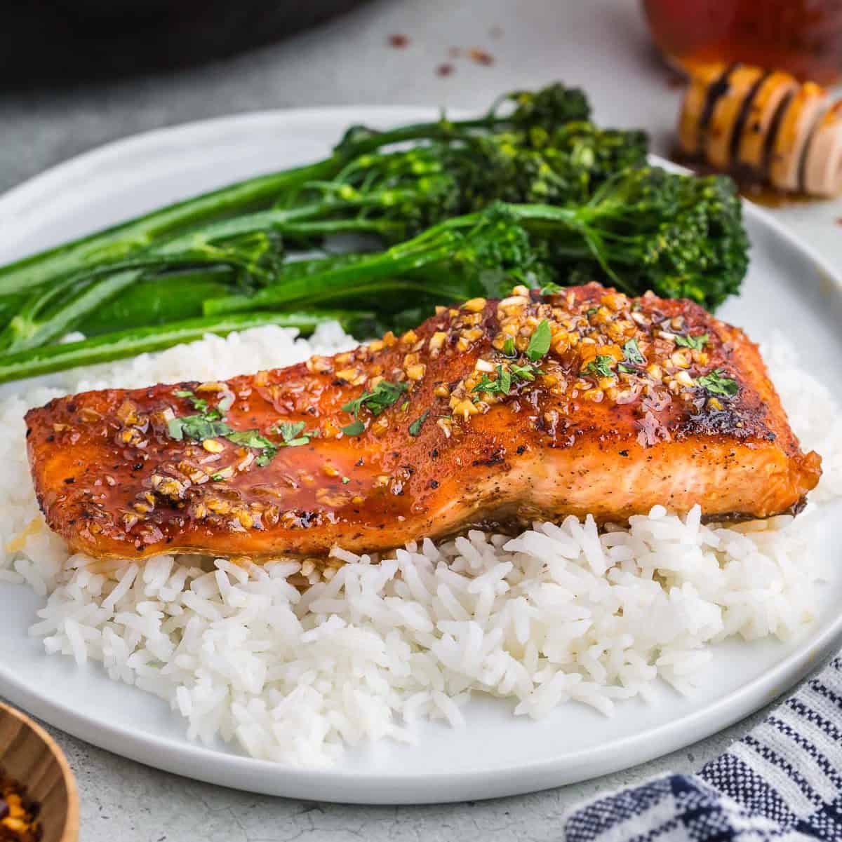 Side shot of a fillet of hot honey salmon sitting atop of a bed of white rice with a side of broccolini all on a white ceramic plate. The hot honey salmon has a golden crispy sear on the flesh with small chunks of garlic and a golden brown hot honey glaze, garnished with fresh parsley.