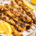 Skewers of garlic chicken parmesan laid out on a white plate garnished with lemon slices and freshly chopped parmesan. Fresh ingredients are peeking-in to the shot in the corners like lemon, parsley, parmesan block, and a brown bowl of onions.