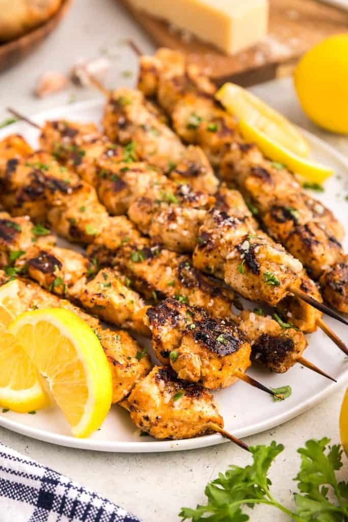 Skewers of garlic chicken parmesan laid out on a white plate garnished with lemon slices and freshly chopped parmesan. Fresh ingredients are peeking-in to the shot in the corners like lemon, parsley, parmesan block, and a brown bowl of onions.