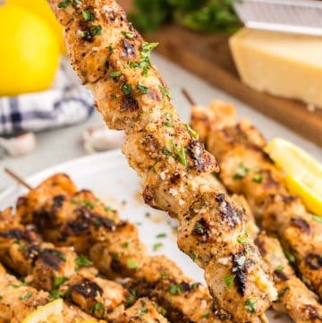 Close up shot of a garlic parmesan chicken skewer being pick up off the serving plate. It is covered in a glistening butter sauce with bits of parmesan, and it is garnished with freshly chopped parsley.