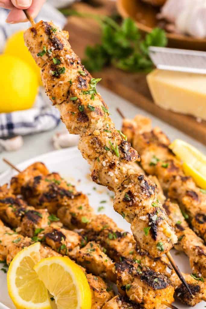Close up shot of a garlic parmesan chicken skewer being pick up off the serving plate. It is covered in a glistening butter sauce with bits of parmesan, and it is garnished with freshly chopped parsley.