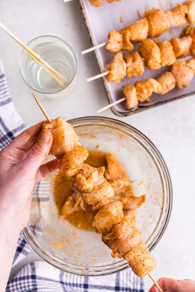 Overhead shot of a bamboo skewer filled with marinated chicken chunks.. In the top right corner of the shot is a metal pan with other chicken skewers ready to go. In the left hand corner is a large glass of water with a bamboo skewer hydrating in it.
