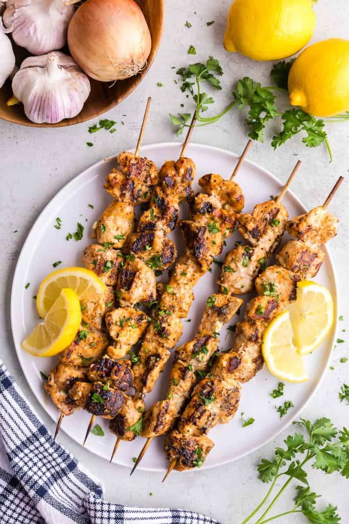 Overhead shot of garlic parmesan chicken skewers served on a white plate with lemon slices and freshly chopped parsley for garnish. The top left hand corner has a wooden bowl filled with fresh onion and garlic peeking into the scene. In the top right hand corner there are a couple of fresh lemons and a spring of parsley.