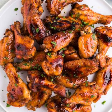 Close up overhead shot of a white plate filled with golden brown crispy hot honey chicken wings garnished with fresh chopped parsley. In the corners of the photo peeking in are a jar of hot honey, a pinch bowl of chili pepper flakes, a bulb of garlic, and a blue linen tea towel.
