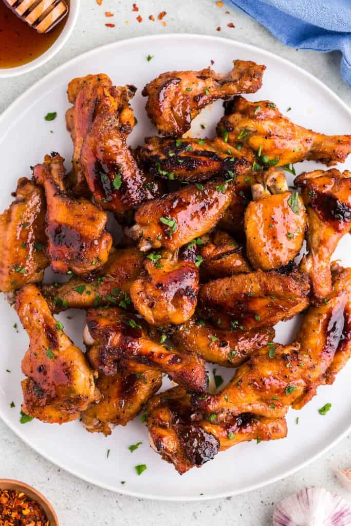 Close up overhead shot of a white plate filled with golden brown crispy hot honey chicken wings garnished with fresh chopped parsley.  In the corners of the photo peeking in are a jar of hot honey, a pinch bowl of chili pepper flakes, a bulb of garlic, and a blue linen tea towel.