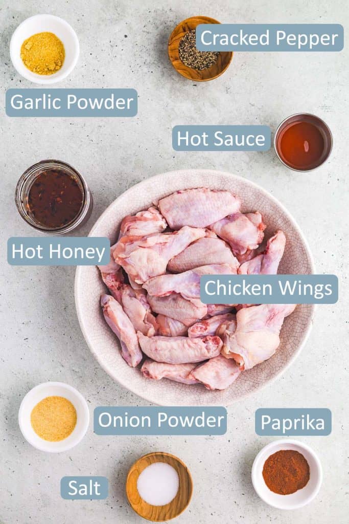 Overhead shot of all the ingredients needed to make hot honey chicken wings.  Ingredients include: Chicken wings, garlic powder, onion powder, black pepper, hot sauce, hot honey, salt, and paprika