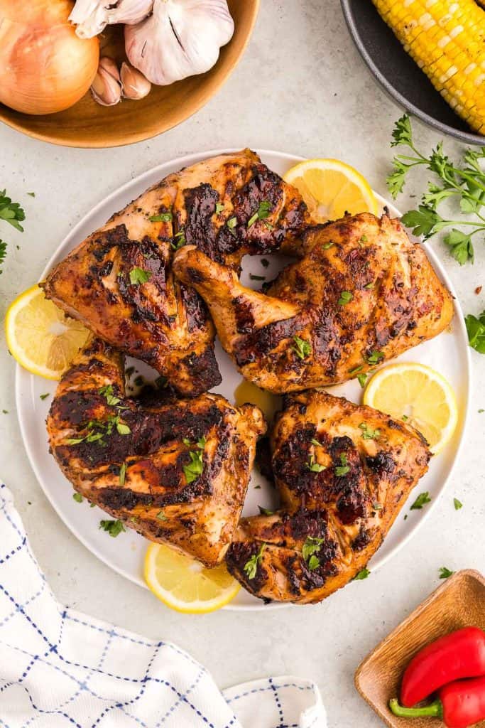 Overhead shot of a white serving plate filled with grilled Portuguese Chicken, and garnished with lemon slices and freshly chopped parsley.  In the right top corner is a bowl of corn on the cobb peeking in.  The top left is a wooden bowl of fresh onions and garlic bulbs.  The bottom right corner has a small wooden bowl with a few red Fresno chilis.