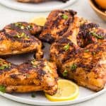 A white serving plate filled with golden brown peri peri chicken with grill marks. The plate is garnished with lemon slices and freshly chopped parsley. Some red fresno peppers, onion, and garlic bulbs are peeking in to the corners of the scene.