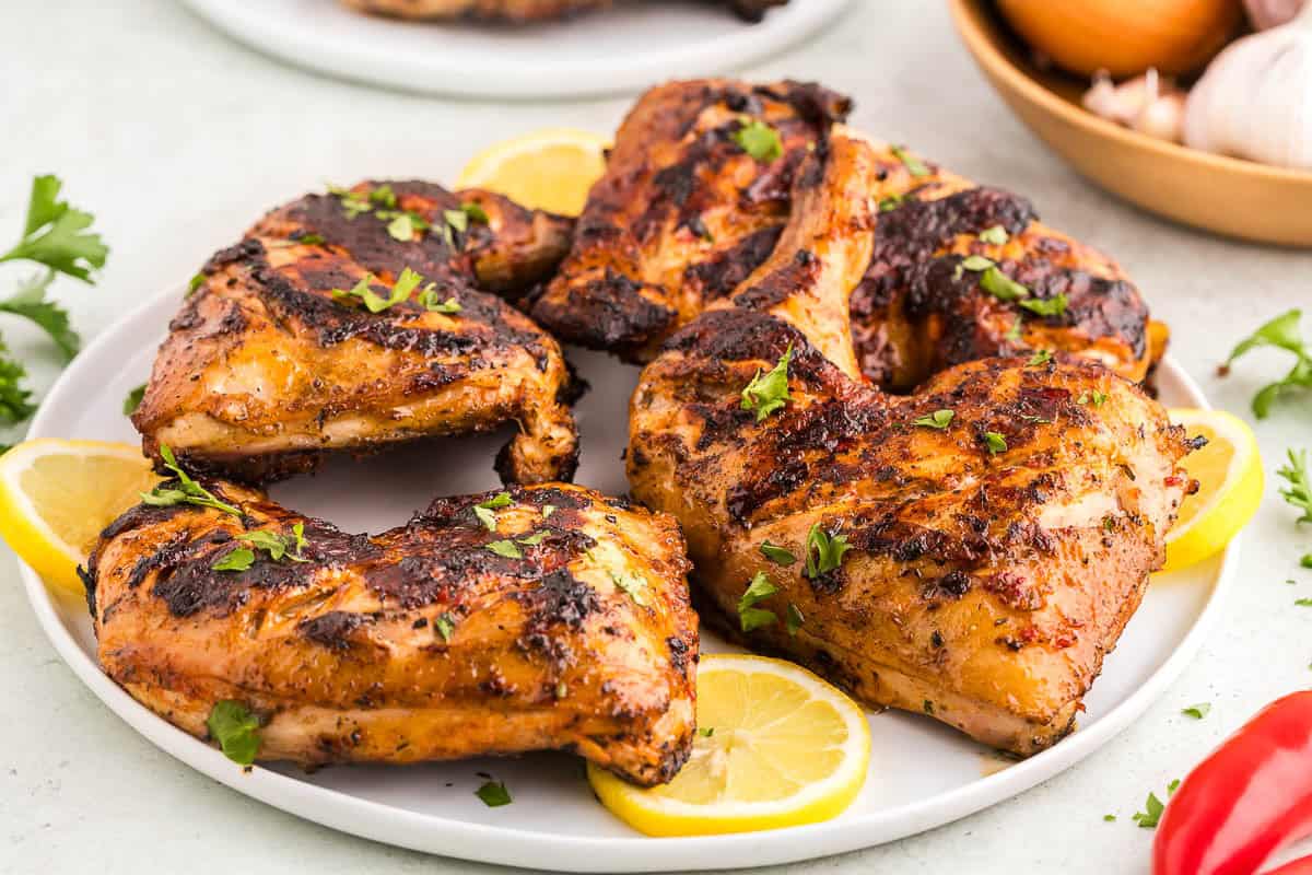 A white serving plate filled with golden brown peri peri chicken with grill marks. The plate is garnished with lemon slices and freshly chopped parsley. Some red fresno peppers, onion, and garlic bulbs are peeking in to the corners of the scene.