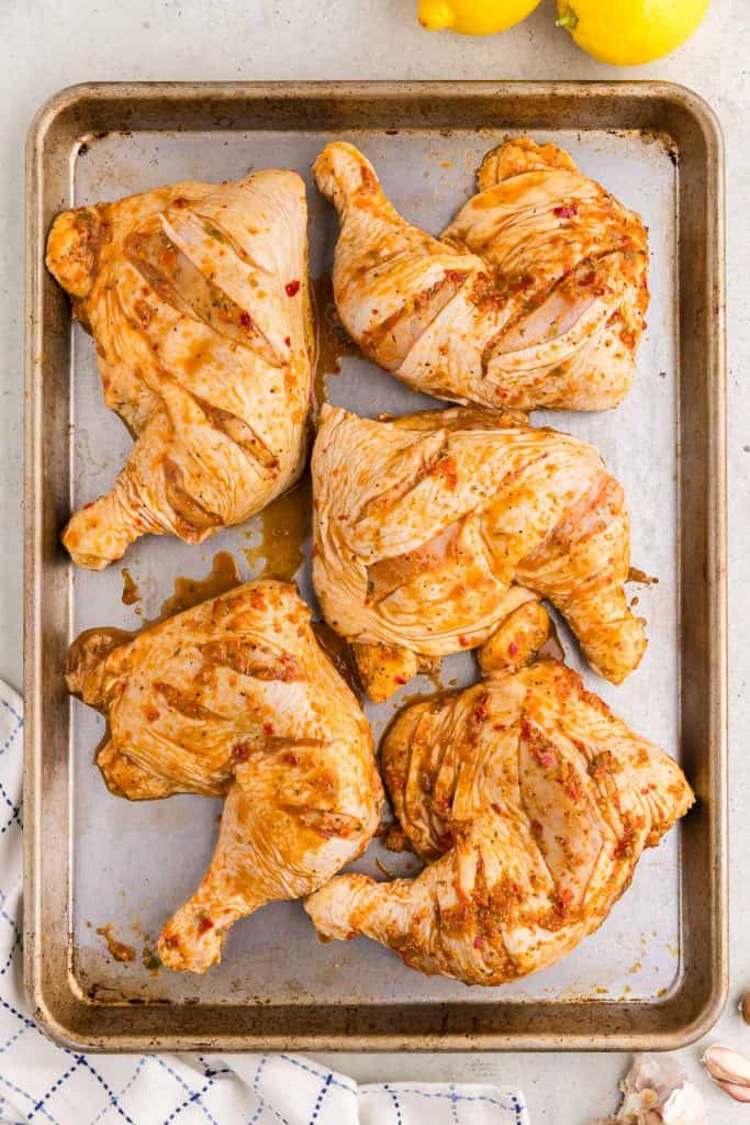 Chicken quarters that have been marinated overnight to make Portuguese Chicken.  The chicken quarters are a light orange from the marinade and are sitting on a silver baking sheet ready to be taken out to the grill.