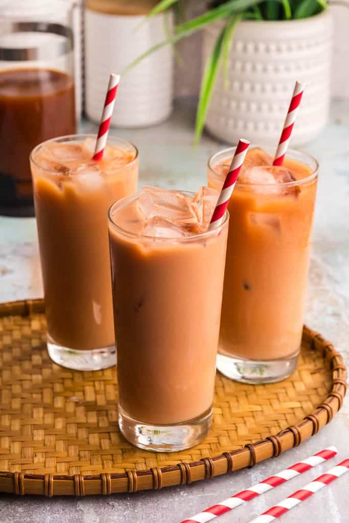 Three tall glasses filled with ice and Thai iced tea sitting on a bamboo serving tray. They each have a red and white straw sticking out of them. In the background the French press with strong black tea is poking in in the left top corner, and in the bottom right corner a few red and white straws are scattered about.