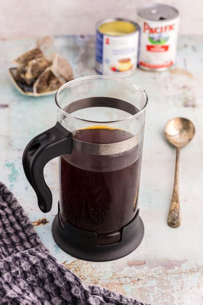 A French press is brewing a very strong black tea. In the background is a pile of the tea bags that were used to brew the tea next to an opened cans of condensed and evaporated milks. A long spoon lays next to the French press.