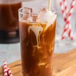 Making a Thai Iced tea. A tall glass filled with ice has a dark tea in it filled almost full. A small ceramic white jug filled with a creamy milk is being poured overtop of the ice and creamy swirls are forming in the dark tea itself.