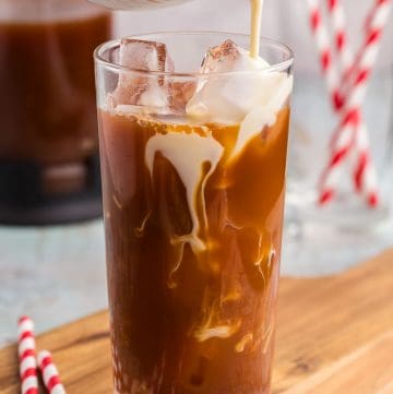 Making a Thai Iced tea. A tall glass filled with ice has a dark tea in it filled almost full. A small ceramic white jug filled with a creamy milk is being poured overtop of the ice and creamy swirls are forming in the dark tea itself.