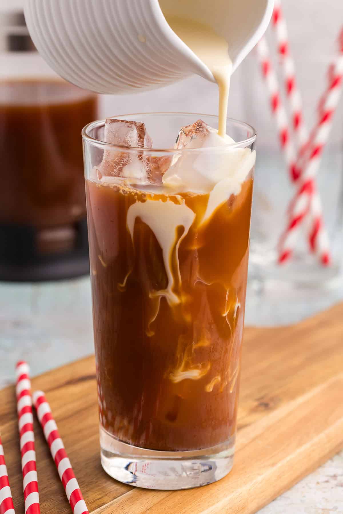 Making a Thai Iced tea. A tall glass filled with ice has a dark tea in it filled almost full. A small ceramic white jug filled with a creamy milk is being poured overtop of the ice and creamy swirls are forming in the dark tea itself.