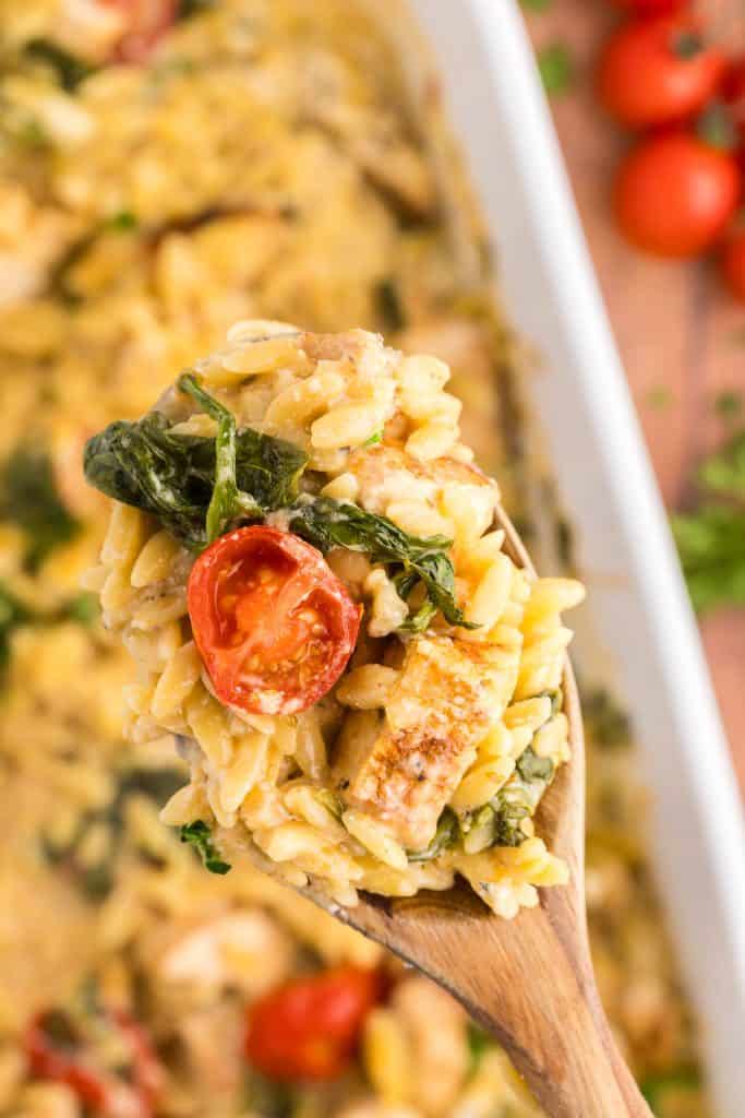 Overhead shot of a wooden serving spoon holding up a spoonful of Chicken Orzo Bake to the camera. The spoon is filled with creamy orzo topped with a halved juicy tomato, a golden brown chunk of chicken and some spinach. In the background below is the casserole dish the serving came out of.