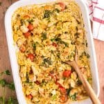 Overhead shot of a 9x13 white casserole dish filled with Chicken Orzo bake. The overall hue is a light creamy yellow with red cherry tomatoes and green spinach dispersed throughout. Chunks of chicken are scattered throughout the orzo pasta. Fresh ingredients are scattered around the casserole dish like parsley, cherry tomatoes, onion and garlic.