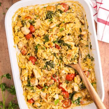 Overhead shot of a 9x13 white casserole dish filled with Chicken Orzo bake. The overall hue is a light creamy yellow with red cherry tomatoes and green spinach dispersed throughout. Chunks of chicken are scattered throughout the orzo pasta. Fresh ingredients are scattered around the casserole dish like parsley, cherry tomatoes, onion and garlic.