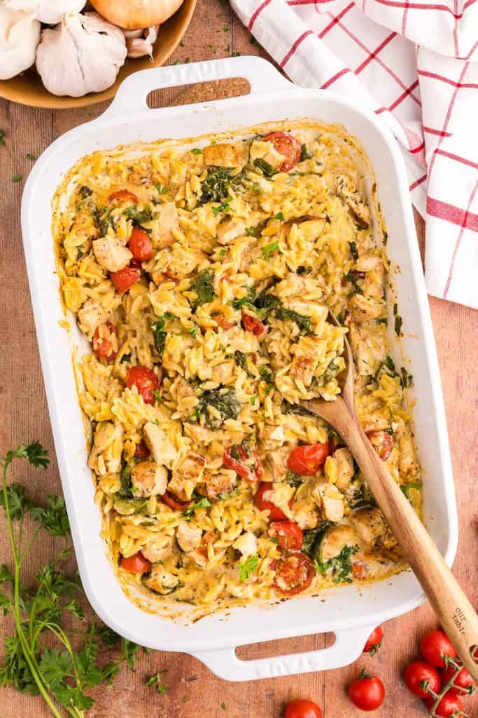 Overhead shot of a 9x13 white casserole dish filled with Chicken Orzo bake. The overall hue is a light creamy yellow with red cherry tomatoes and green spinach dispersed throughout. Chunks of chicken are scattered throughout the orzo pasta. Fresh ingredients are scattered around the casserole dish like parsley, cherry tomatoes, onion and garlic.