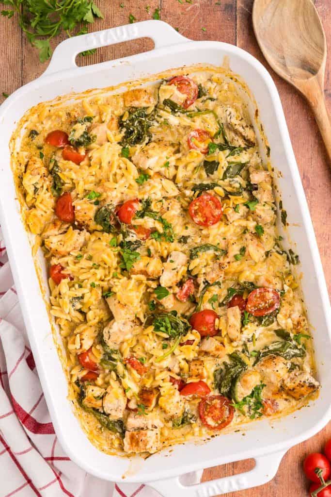 Overhead shot of a white ceramic 9x13 casserole dish set slightly askew. Inside is the baked Orzo Chicken bake. Large gold chunks of chicken in a golden creamy sauce with orzo, wilted spinach, and bright red cherry tomatoes. The top right corner has a wooden serving spoon poking in to the scene. The bottom right has some cherry tomatoes, the top left has a few sprigs of fresh parsley and the bottom left corner has a white linen with red stripes.