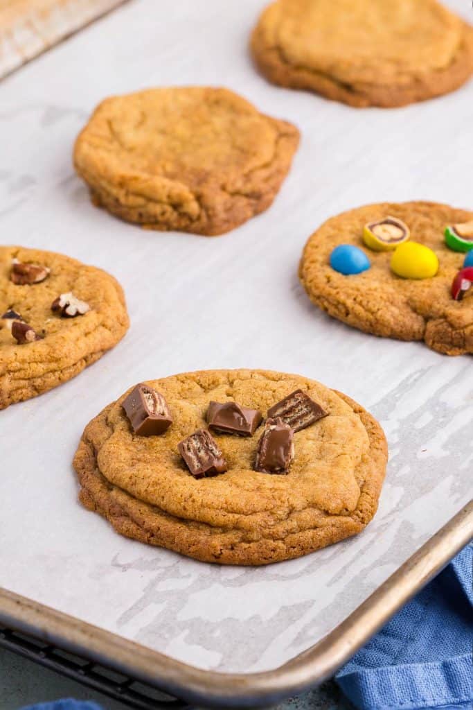 Chocolate chipless cookies on a sheet pan just after coming out of the oven. The closest cookie has had bits of Kit Kat added to the surface, another has chopped up peanut M&Ms and a third has chopped pecans. The rest are plain.