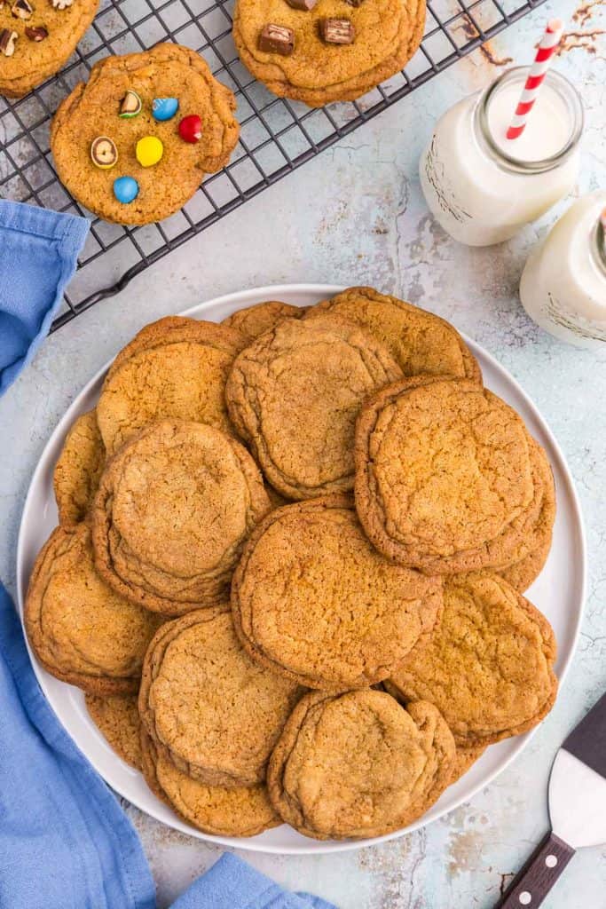 Overhead shot of a large plate of golden brown chocolate chipless cookies. It's chocolate chip cookies without the chips! Above the plate of cookies is a cooling rack with more cookies, but one has M&Ms in it, another with pecans, and one with kit kat bits. In the top right of the picture are two milk glasses with red and white striped straws.