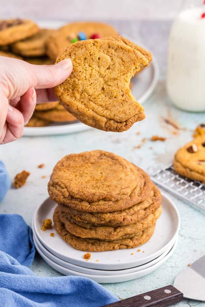 Side shot of a stack of 4 chocolate chipless cookies on a small white plate. A hand is holding another cookie up from the stack and a bite has been taken out of the right hand side. In the background is a large plate filled with more chocolate chipless cookies, a glass of milk with a red and white straw, and just peeking into the frame is a cooling rack with more cookies.