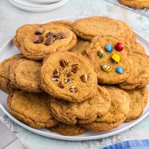 45 degree angle shot of a large plate of chocolate chipless cookies. The plate has mostly plain cookies, but there is also one with chunks of kit kat on the top, another with chopped peanut M&Ms and a third with chopped pecans, to show that this recipe can be tailored to your own tastes.