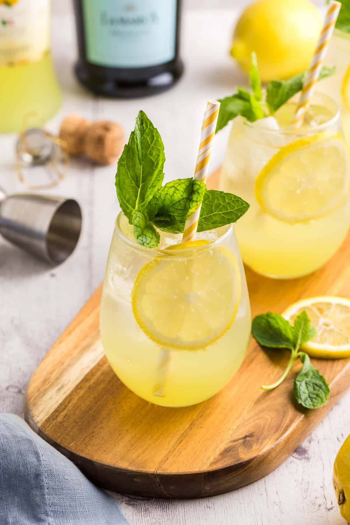 Three limoncello spritz garnished with mint and a yellow straw being served on a long wooden board. In the background are the limoncello and prosecco bottles, and in the foreground in the bottom right a fresh lemon is peeking in to the scene.