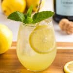 Close up shot of a stemless wine glass sitting on a wood chopping board filled with Limoncello Spritz. The glass is filled with ice and the drink is a yellow ombre, darker yellow on the bottom which lightens as it goes up. There is a yellow straw in the glass and it is garnished with fresh mint.