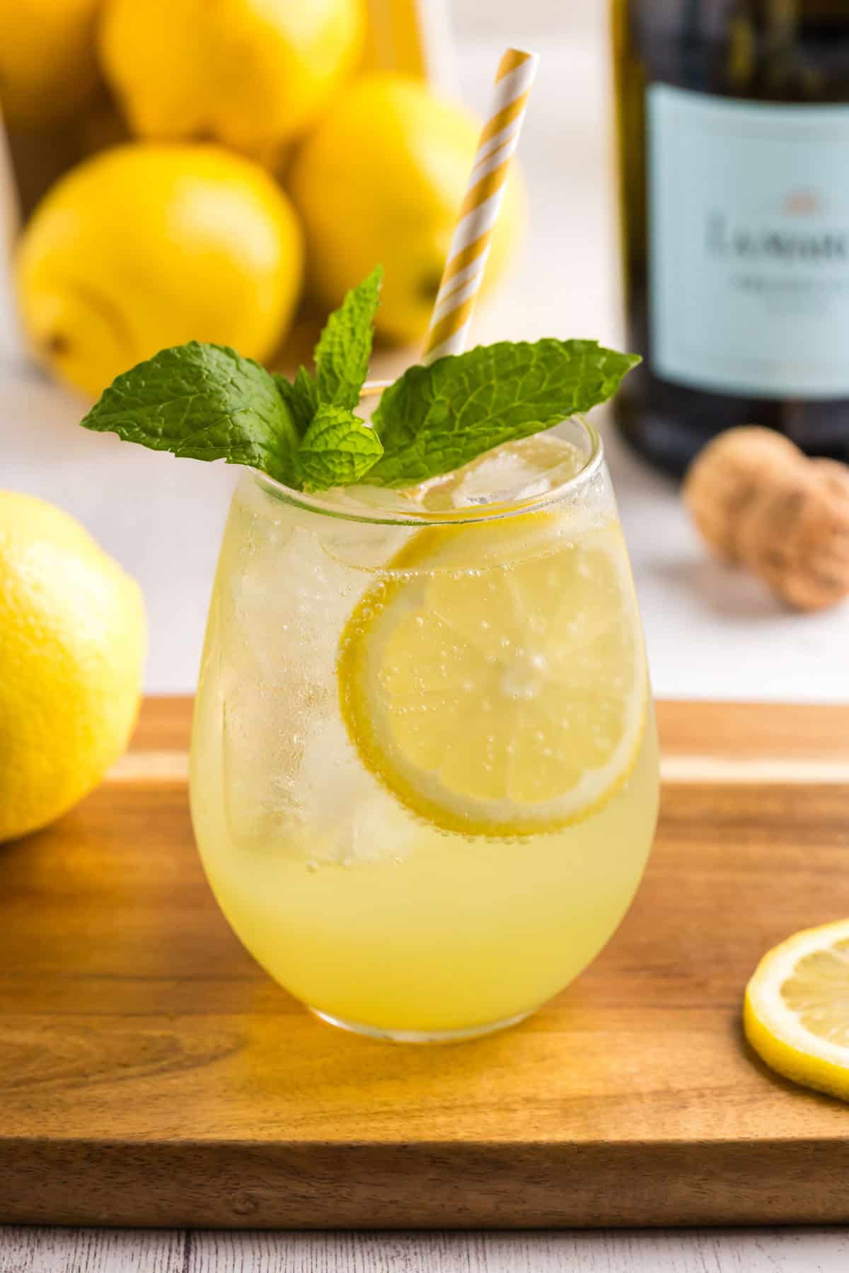 Close up shot of a stemless wine glass sitting on a wood chopping board filled with Limoncello Spritz. The glass is filled with ice and the drink is a yellow ombre, darker yellow on the bottom which lightens as it goes up. There is a yellow straw in the glass and it is garnished with fresh mint.