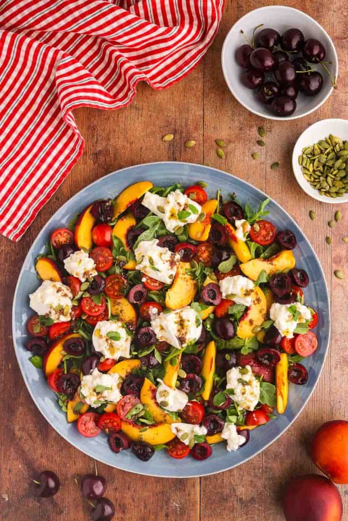 A large platter of peach burrata salad takes up the lower 2/3rds of the photo with some cherries in the lower left corner, peaches in the lower right corner. The upper right corner has a small white bowl filled with cherries and a small pinch bowl of pepitas.