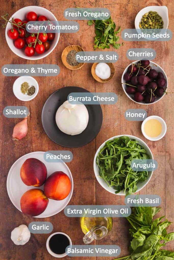 Ingredients needed for peach burrata salad laid out on a wooden table. Ingredients include: arugula, fresh basil and oregano, fresh cherries, peaches, honey, balsamic vinegar, burrata cheese, salt and pepper, dried oregano and thyme, shallot, extra virgin olive oil, cherry tomatoes, pumpkin seeds, fresh garlic