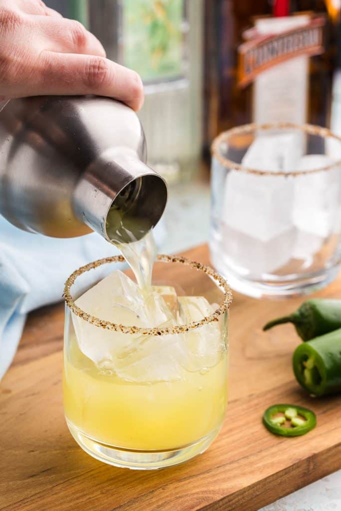 a 45 degree angle shot of a cocktail shaker pouring a pineapple jalapeno margarita into a tajin-sugar rimmed low ball glass filled with ice. The glass is 3/4 full of a soft yellow liquid, while more margarita is pouring out of the cocktail shaker. In the background is another rimmed glass filled with ice waiting to be filled. Some green sliced jalapenos are in the foreground.