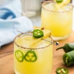 Side angle shot of a low ball glass filled with ice and a gentle yellow pineapple jalapeno margarita. The rim of the glass is rimmed with tajin and sugar. There is a cocktail pick running across the top of the glass with a slice of pineapple and a thin slice of jalapeno on top. In the glass there are two slices of jalapeno floating between the ice and the side of the glass.