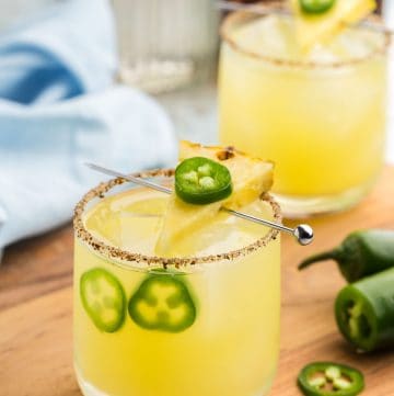 Side angle shot of a low ball glass filled with ice and a gentle yellow pineapple jalapeno margarita. The rim of the glass is rimmed with tajin and sugar. There is a cocktail pick running across the top of the glass with a slice of pineapple and a thin slice of jalapeno on top. In the glass there are two slices of jalapeno floating between the ice and the side of the glass.
