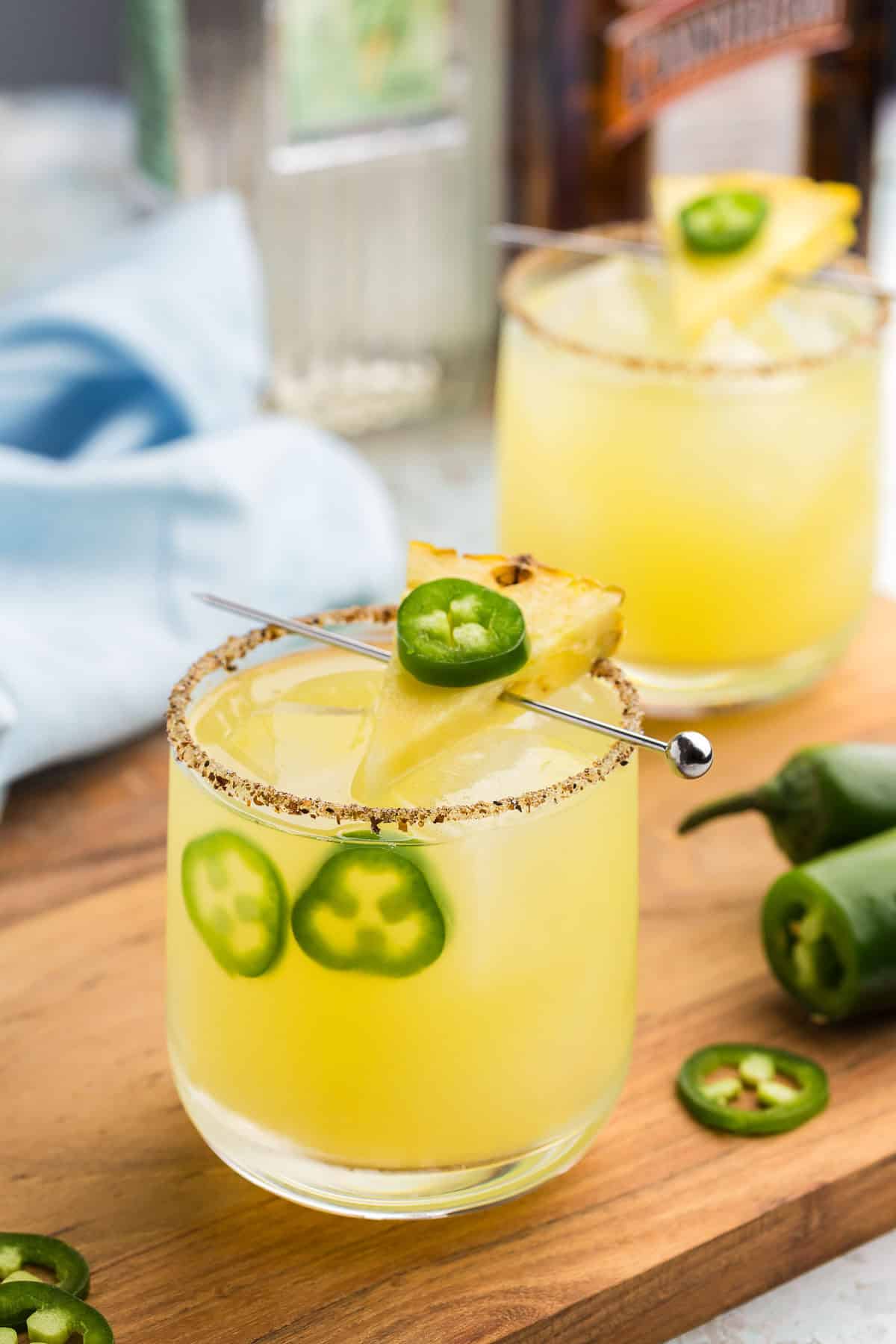 Side angle shot of a low ball glass filled with ice and a gentle yellow pineapple jalapeno margarita. The rim of the glass is rimmed with tajin and sugar. There is a cocktail pick running across the top of the glass with a slice of pineapple and a thin slice of jalapeno on top. In the glass there are two slices of jalapeno floating between the ice and the side of the glass.