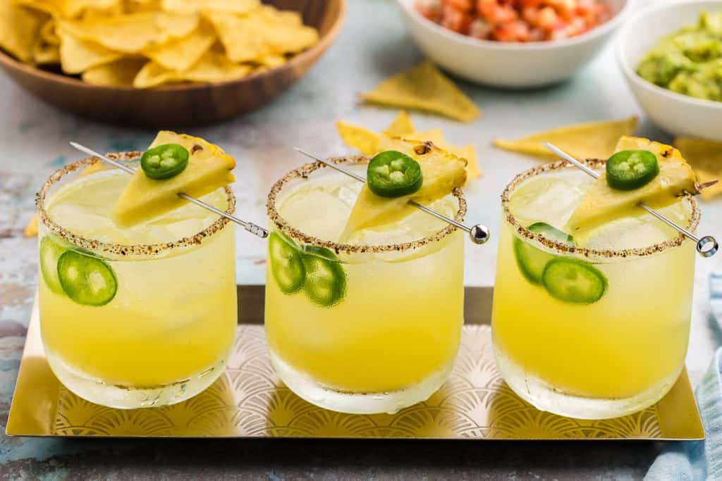 A side shot of three pineapple jalapeno margaritas in low ball glasses. Each one is a soft sunshine yellow with a wedge of pineapple skewered on a cocktail skewer with a round slice of green jalapeno on top. Two round jalapeno slices are also floating in each glass between the ice and the wall of the glass. Behind the margaritas is a wooden bowl filled with yellow tortilla chips, and two small white bowls, one with red salsa and the other with green guacamole.