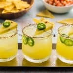 horizontal shot at a shallow 45 angle. Three low ball glasses sitting on a narrow silver tray in a straightline. Each glass has a tajin sugar rim, is filled with ice, and is filled with a soft yellow pineapple jalapeno margarita. Each glass has a cocktail pick with a pineapple wedge and jalapeno slice sitting on top. There is also a couple of jalapeno round slices floating in each drink between the ice and the edge of the glass.
