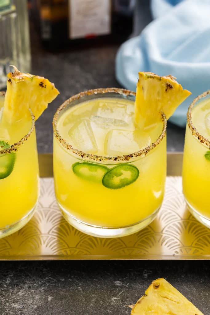 Three pineapple jalapeno margaritas on a serving tray. They are bright yellow, have a few slices of green jalapeno in them and a pineapple wedge on the rim. The glass has been rimmed with Tajin.