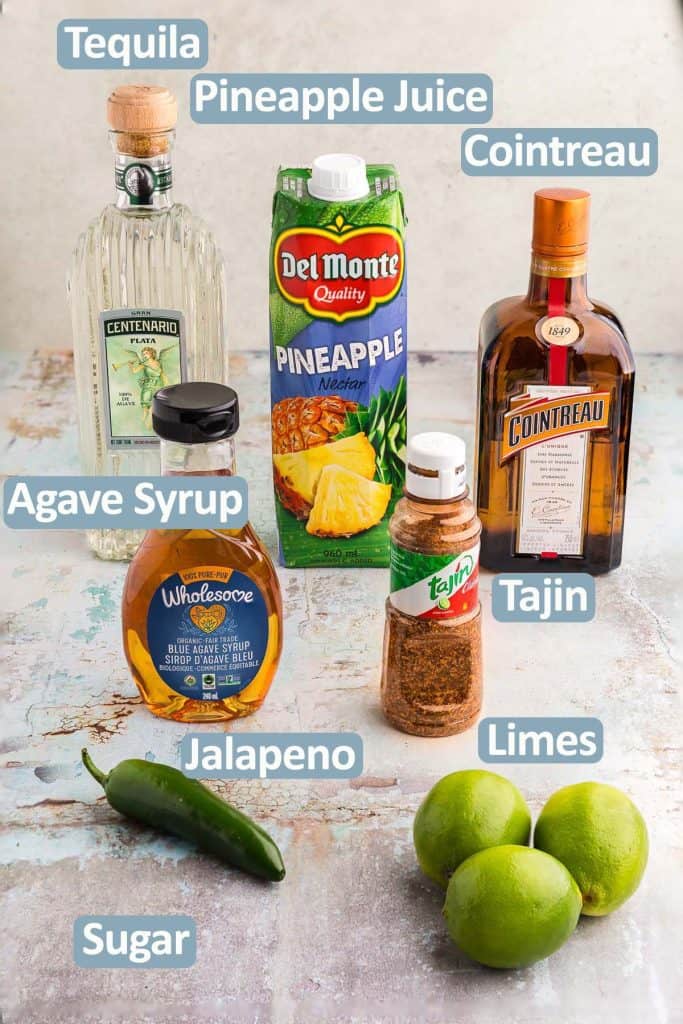 45 degree angle shot of the ingredients needed to make a pineapple jalapeno margarita. Ingredients include: Tequila, Pineapple juice, Cointreau, Agave syrup, Tajin, Fresh Jalapeno, Fresh limes, and white sugar.