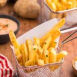 Close up side shot of a small metal basket lined with parchment paper and filled with Air fryer frozen french fries. The fries have been seasoned with salt, pepper, and fresh parsley. In th background are some potates, fry sauce, and another small basket of fries.