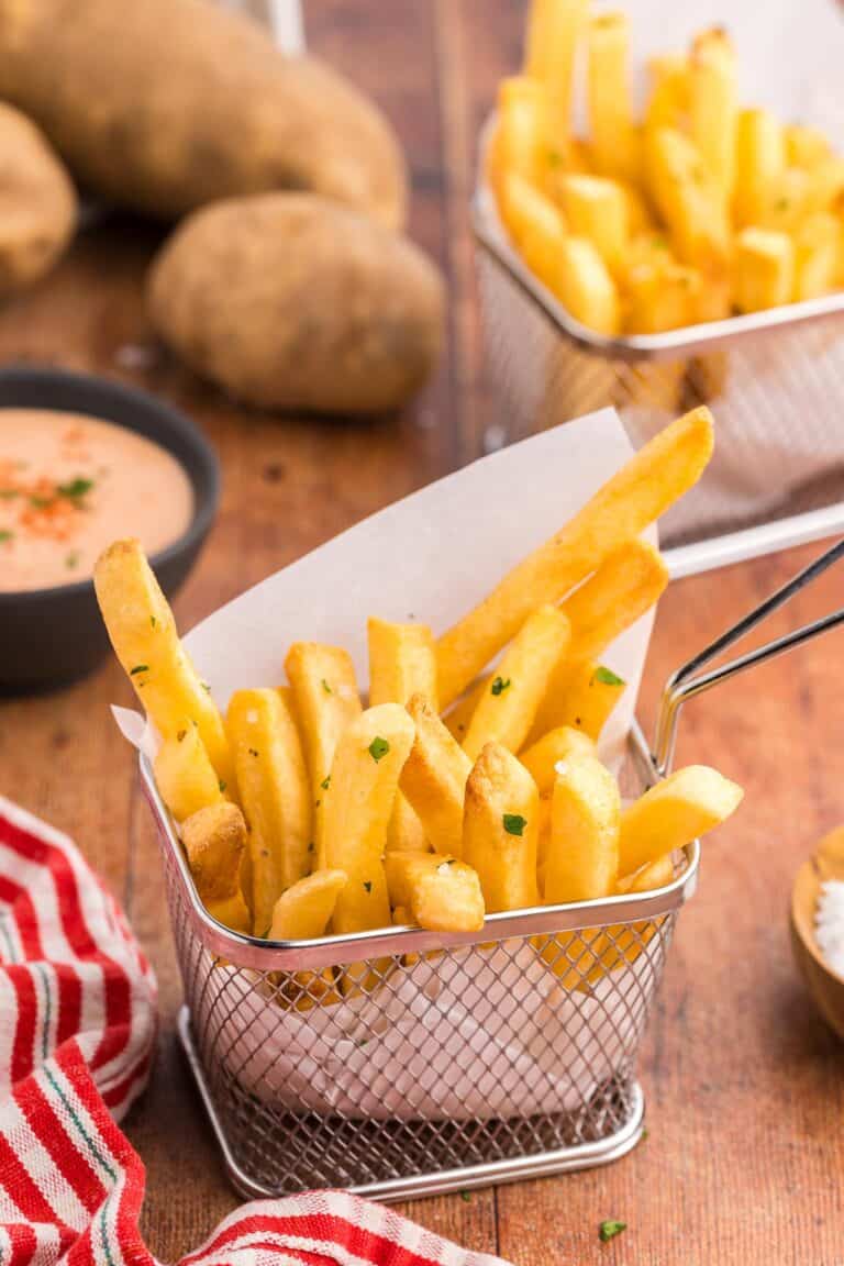 Close up side shot of a small metal basket lined with parchment paper and filled with Air fryer frozen french fries. The fries have been seasoned with salt, pepper, and fresh parsley. In th background are some potates, fry sauce, and another small basket of fries.
