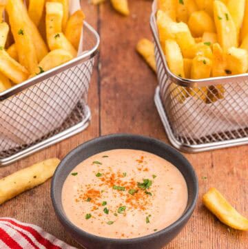45 degree angle shot of a small dark gray bowl filled with an orangey/pink sauce called French Fry Sauce. The sauce has paprika sprinkled over top and a little bit of freshly chopped parsley. There are two small baskets of fries on either side of the bowl.
