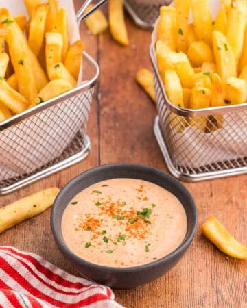45 degree angle shot of a small dark gray bowl filled with an orangey/pink sauce called French Fry Sauce. The sauce has paprika sprinkled over top and a little bit of freshly chopped parsley. There are two small baskets of fries on either side of the bowl.