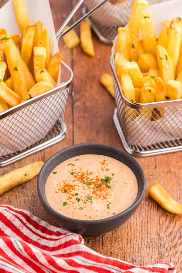 45 degree angle shot of a small dark gray bowl filled with an orangey/pink sauce called French Fry Sauce. The sauce has paprika sprinkled over top and a little bit of freshly chopped parsley. There are two small baskets of fries on either side of the bowl.