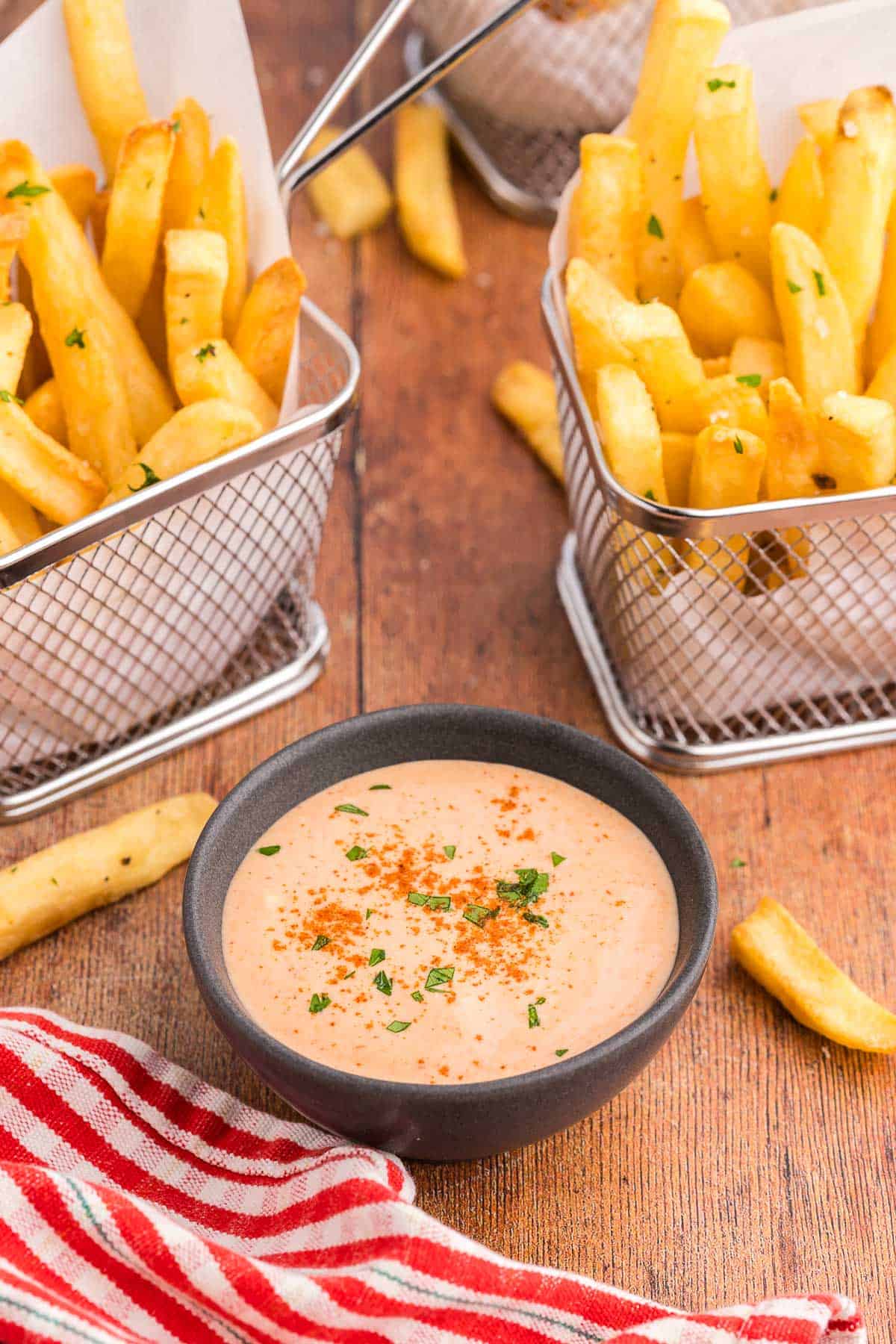 45 degree angle shot of a small dark gray bowl filled with an orangey/pink sauce called French Fry Sauce. The sauce has paprika sprinkled over top and a little bit of freshly chopped parsley. There are two small baskets of fries on either side of the bowl.