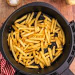 overhead shot of golden brown air fryer frozen French fries sitting in the air fryer basket. Two wooden pinch bowls in the upper left corner contain salt and pepper. In the top right corner is a basket of potatoes and the bottom right corner there is a potato peeking into the scene. The bottom left corner has a red and white check linen.
