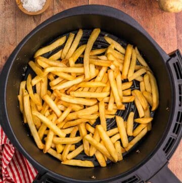 overhead shot of golden brown air fryer frozen French fries sitting in the air fryer basket. Two wooden pinch bowls in the upper left corner contain salt and pepper. In the top right corner is a basket of potatoes and the bottom right corner there is a potato peeking into the scene. The bottom left corner has a red and white check linen.