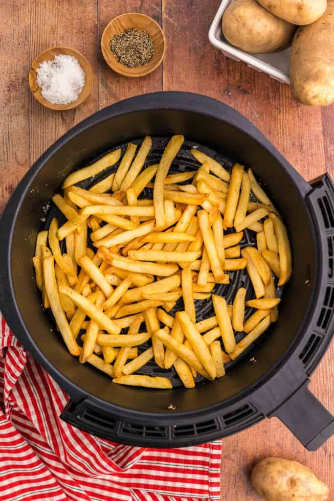 overhead shot of golden brown air fryer frozen French fries sitting in the air fryer basket.  Two wooden pinch bowls in the upper left corner contain salt and pepper.  In the top right corner is a basket of potatoes and the bottom right corner there is a potato peeking into the scene.  The bottom left corner has a red and white check linen.